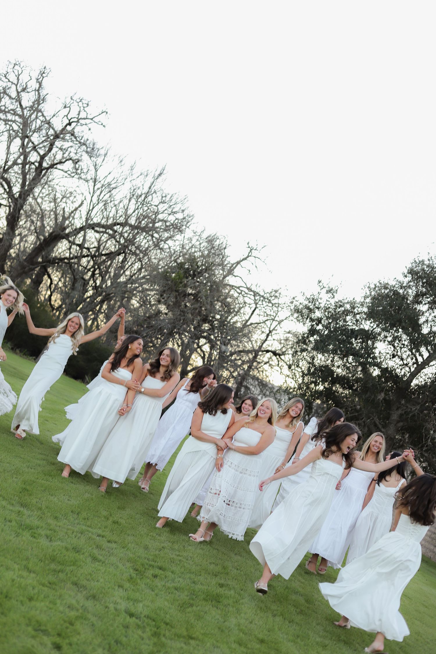 A group of women in matching white dresses pose together on a green lawn with trees in the background.