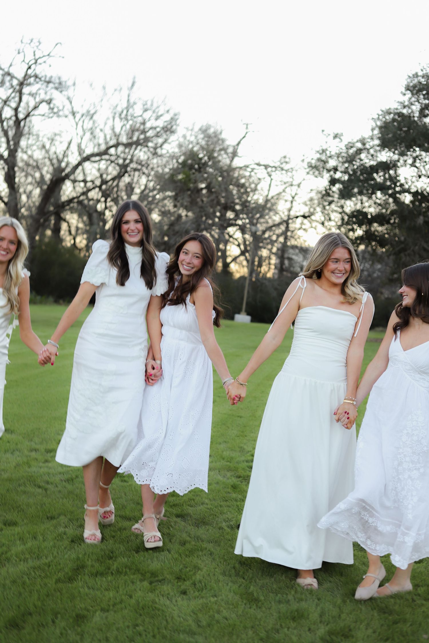 A group of women in white dresses hold hands while walking across a grassy field at dusk.