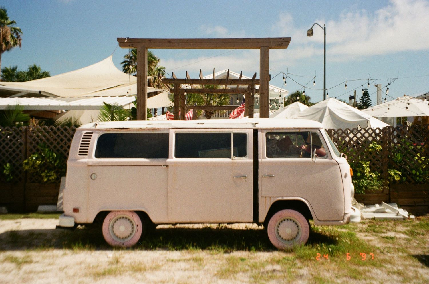 A vintage white Volkswagen bus parked in front of a wooden pergola with palm trees and white canopies in the background.