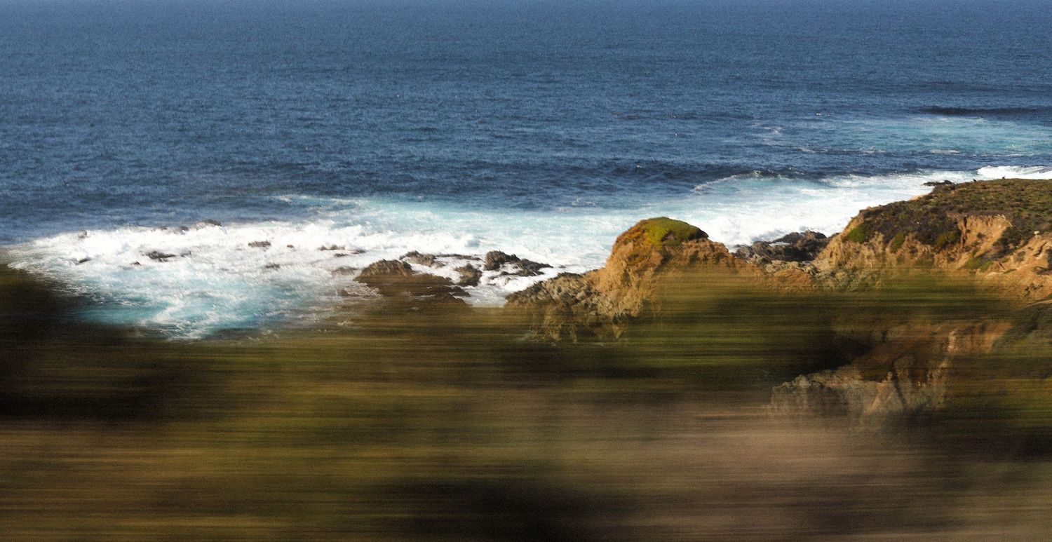 Panoramic view of rocky coastline with crashing waves against blue ocean and cliffs at sunset.