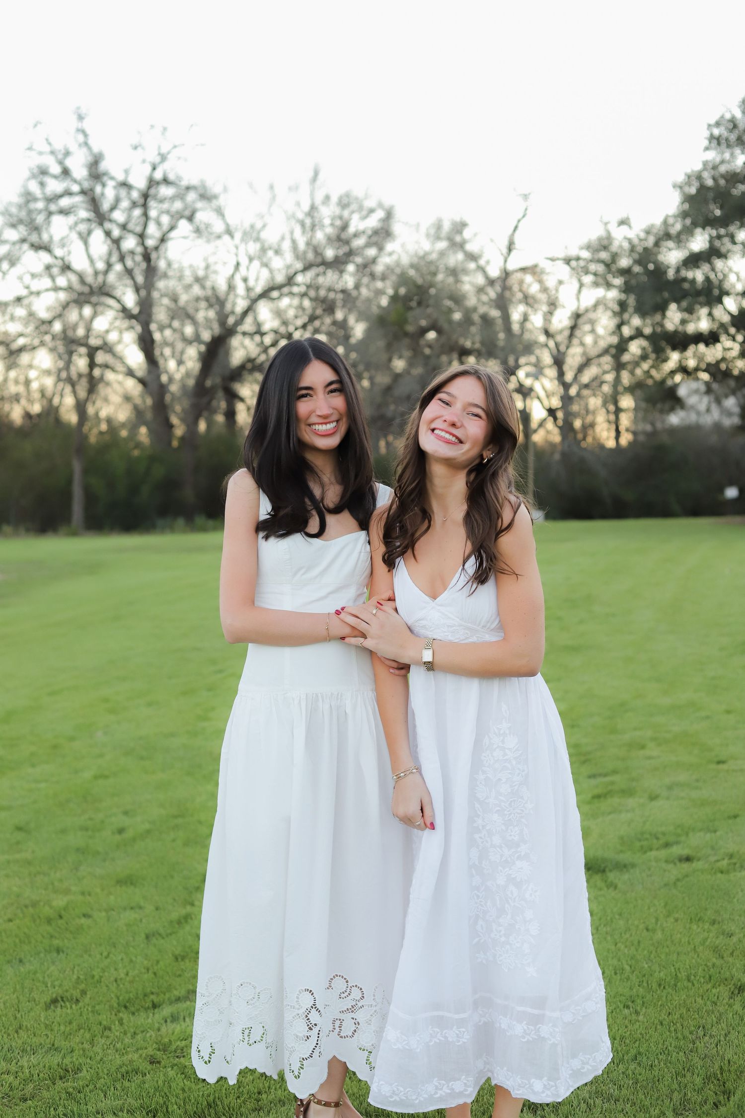 Two friends in white lace dresses pose together on green lawn at sunset.