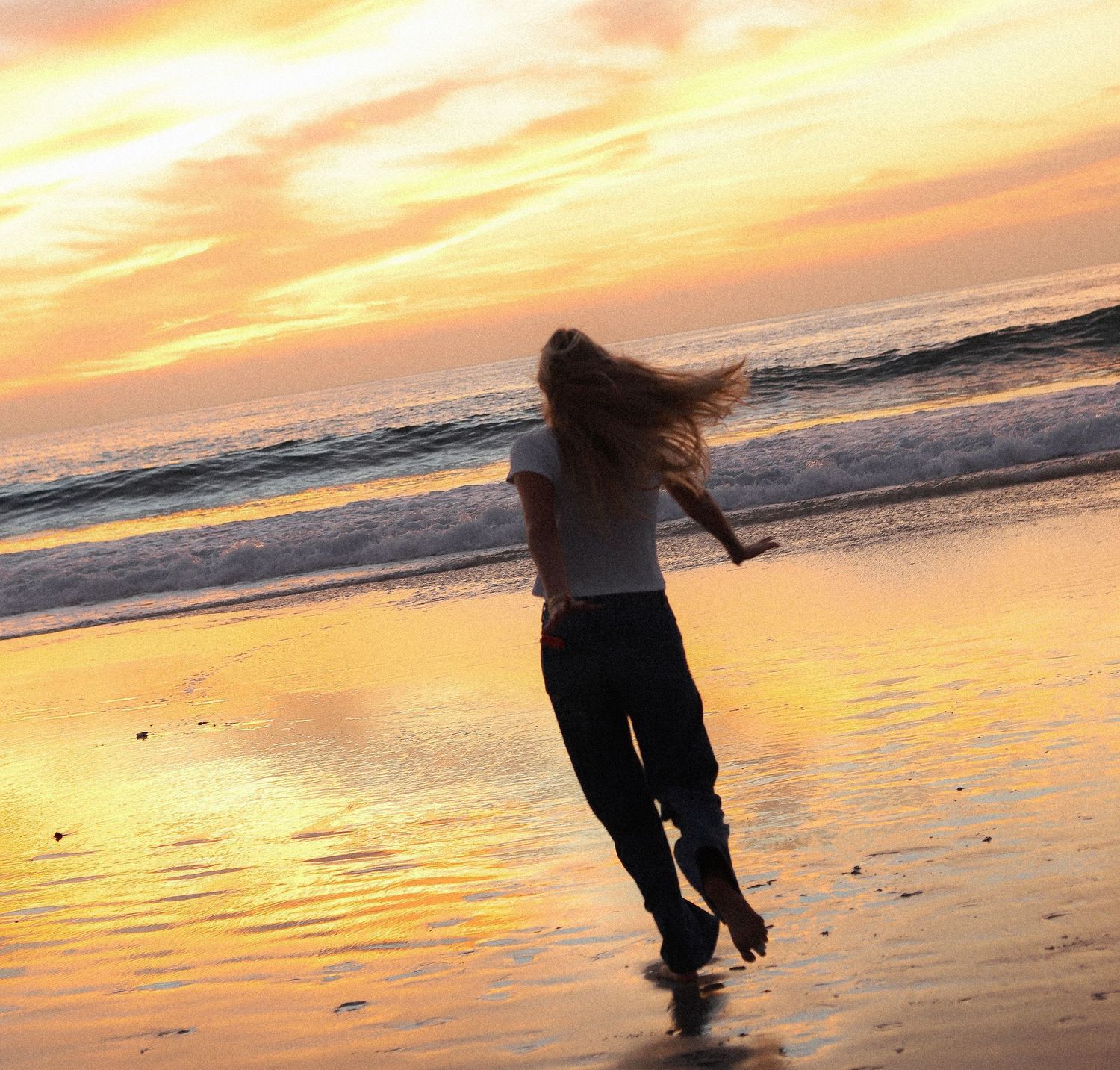 A silhouette dances on a beach at sunset with golden orange sky reflecting on wet sand and ocean waves.