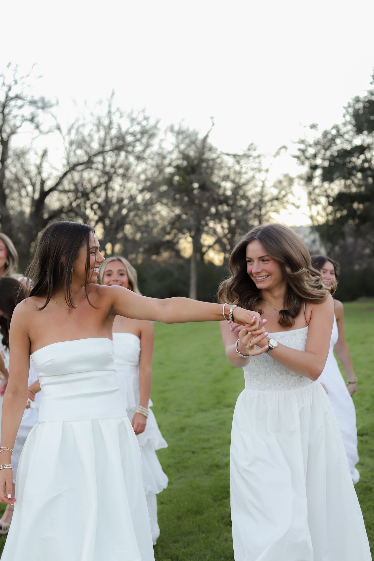 Women in flowing white dresses dance and twirl together on green lawn.