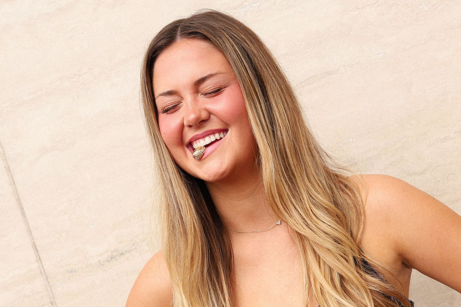 A candid portrait of a woman laughing with her eyes closed against a beige wall.