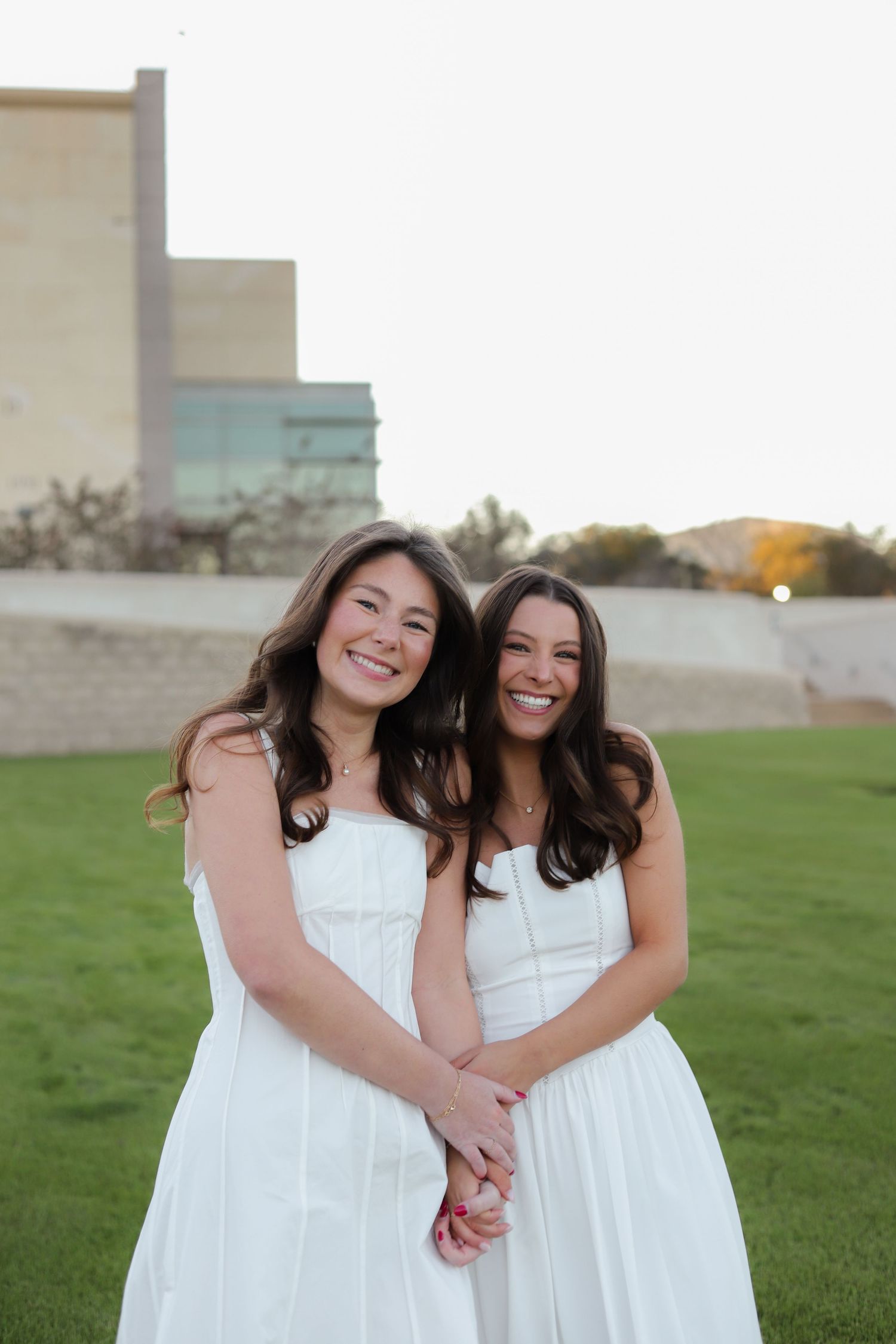 Two friends in matching white dresses smile together while holding hands on a grassy lawn at sunset.