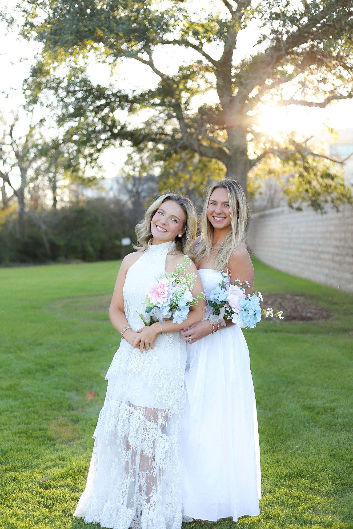 Two women in white dresses pose together holding floral bouquets on a sunny lawn with trees in the background.