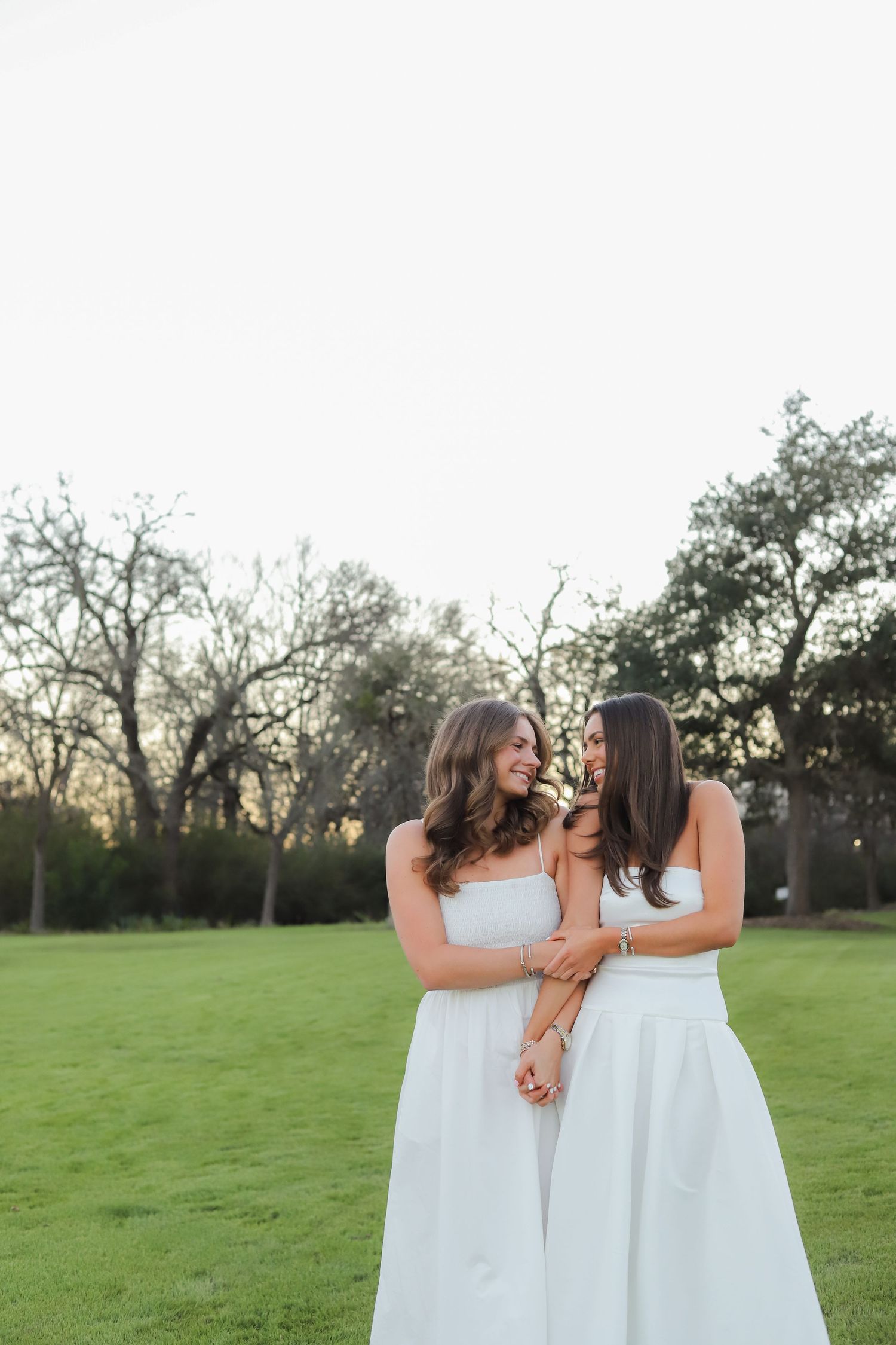 Three women in matching white dresses stand together embracing in a green field at sunset.