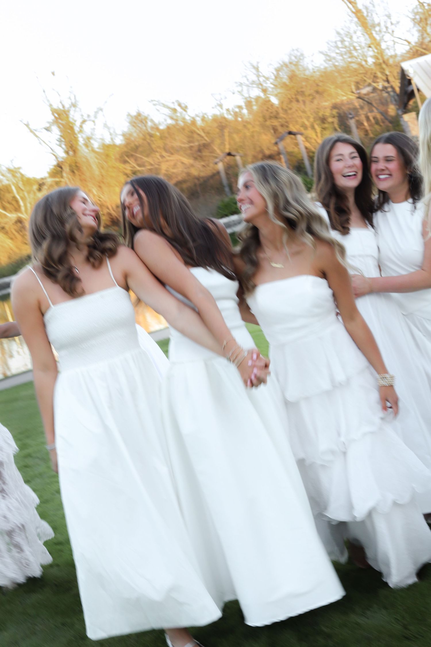 A group of bridesmaids in white dresses laugh together while standing on grass at sunset.