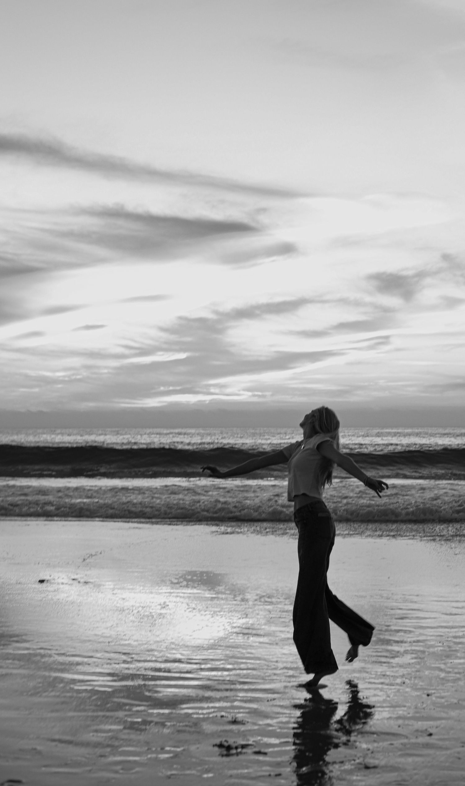 A silhouetted figure dances gracefully along a beach shoreline at sunset in a black and white photograph.