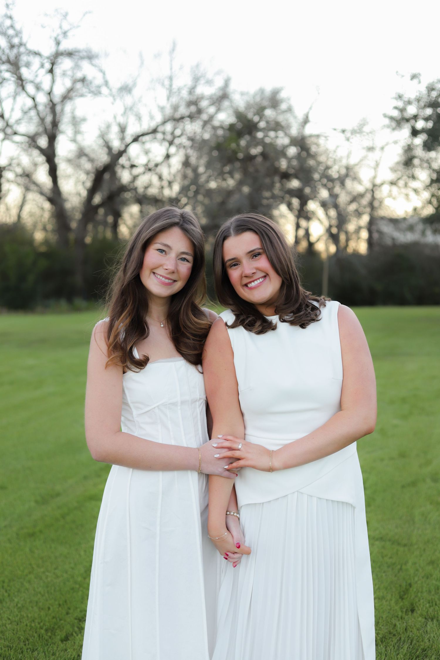 Two women in elegant white dresses pose together on a green lawn during golden hour.