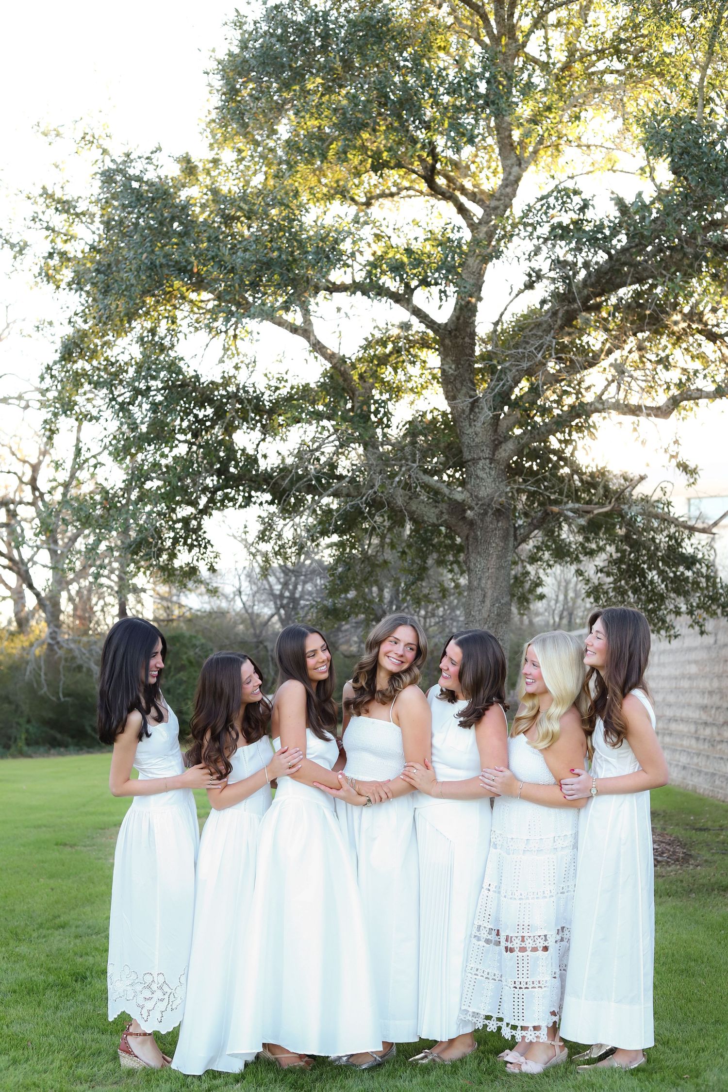 Group of women in white dresses pose together on grassy lawn with trees in background.