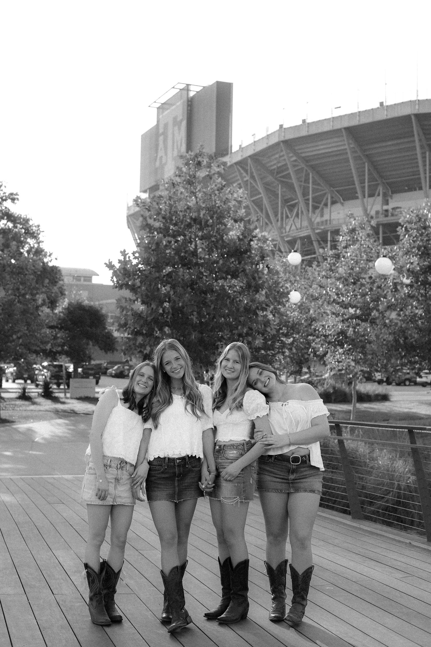 Black and white photo of friends in matching outfits posing on a wooden boardwalk near a stadium.