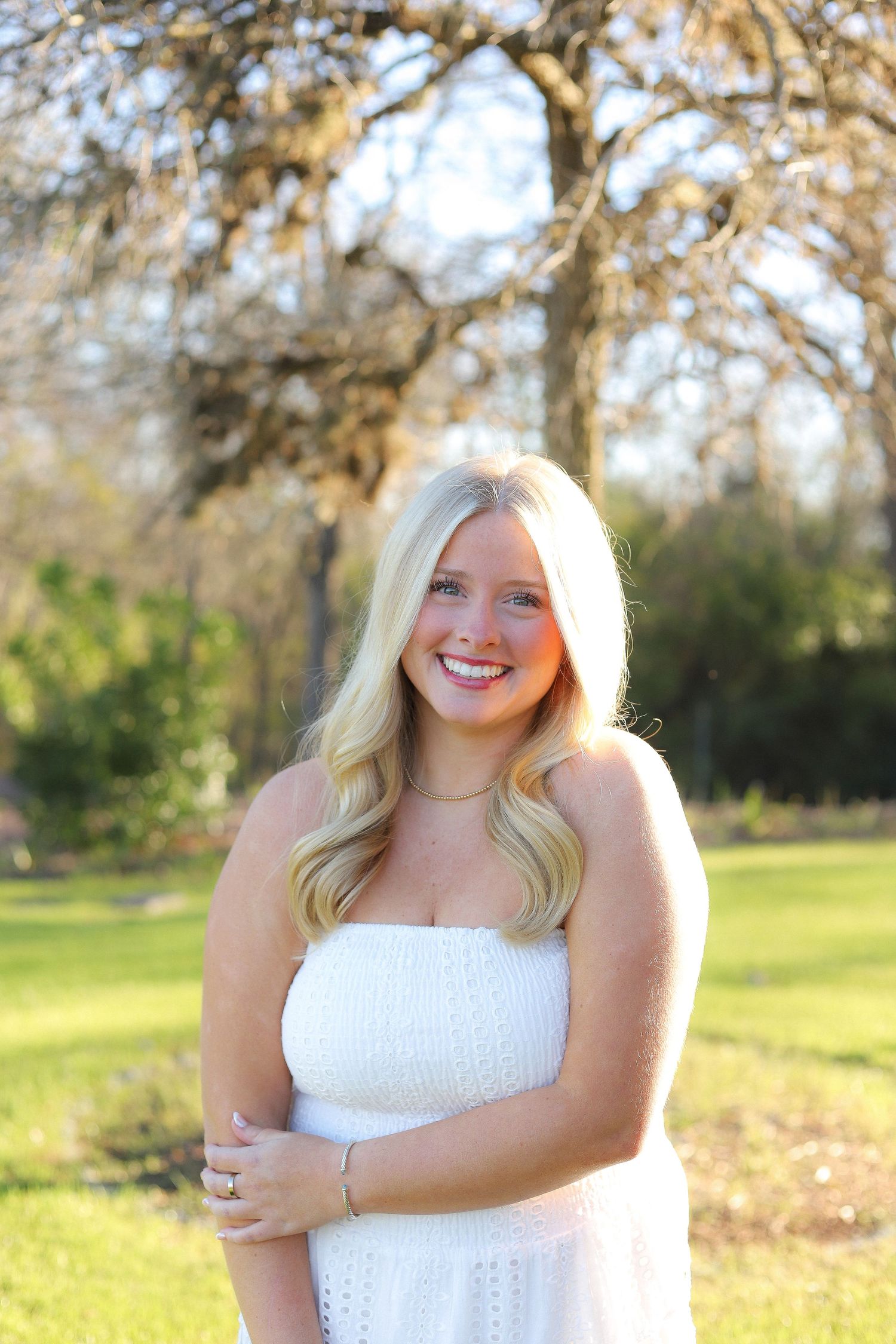 A person wearing a white eyelet dress poses in a grassy field during golden hour.