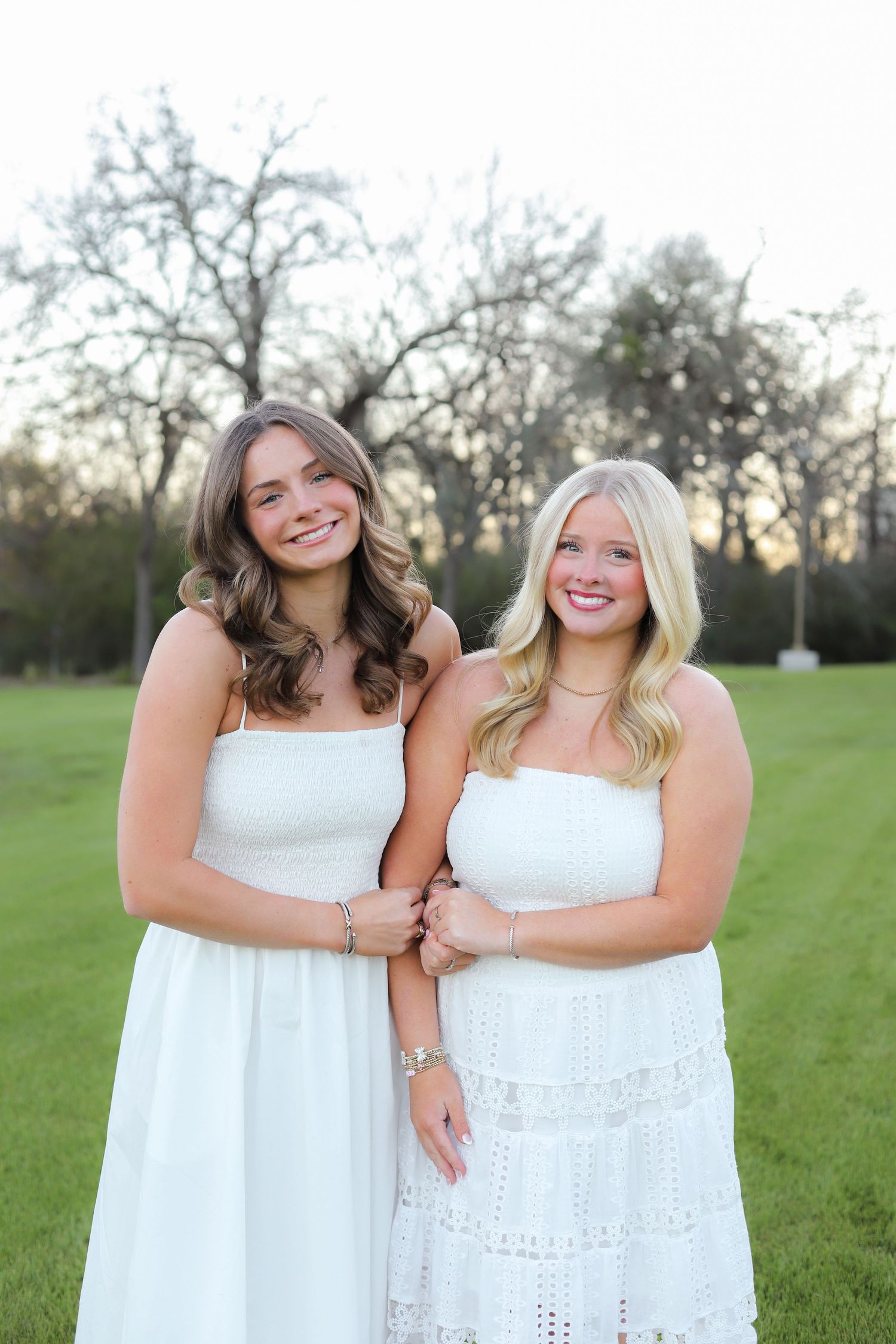 Two friends in white dresses share a candid moment while posing together on a sunny lawn.