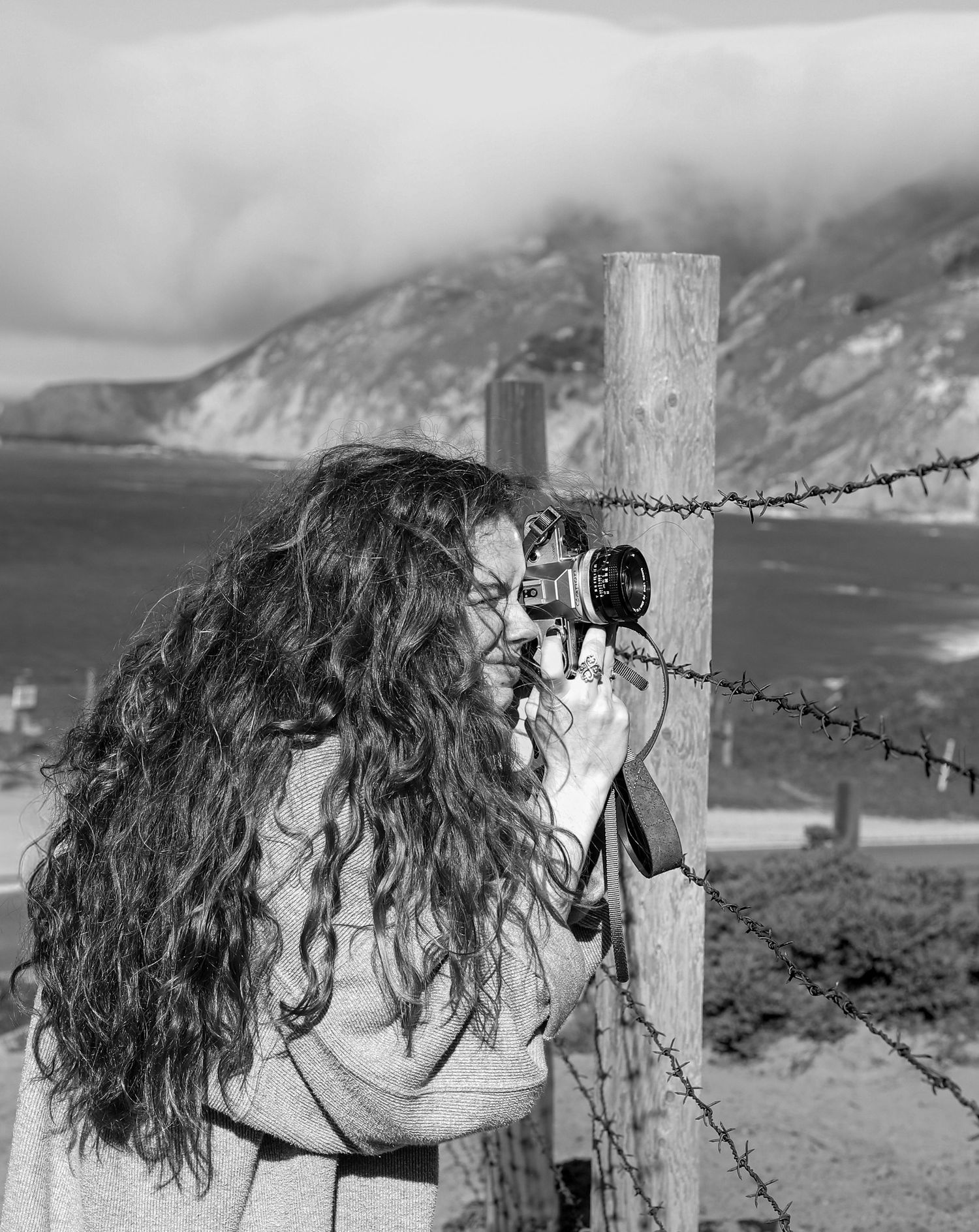 Black and white photo of someone photographing a misty coastal scene through a barbed wire fence.
