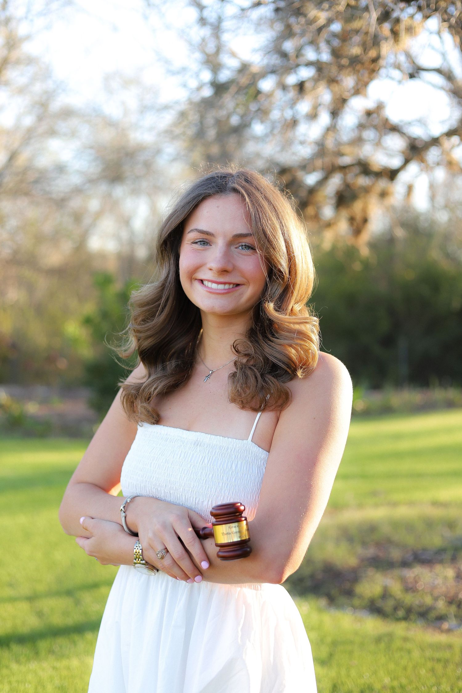 A person in a white dress poses outdoors against a natural backdrop of trees and grass.