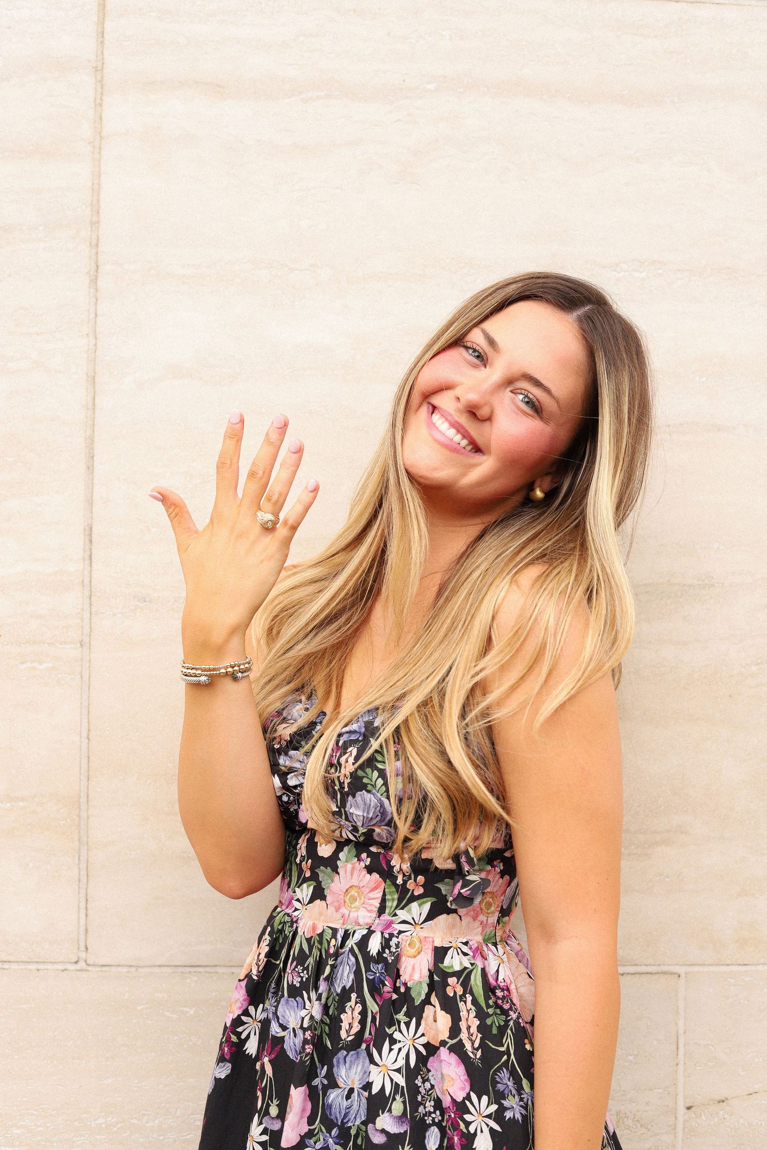 A woman in a floral dress shows off her ring while posing against a light colored wall.