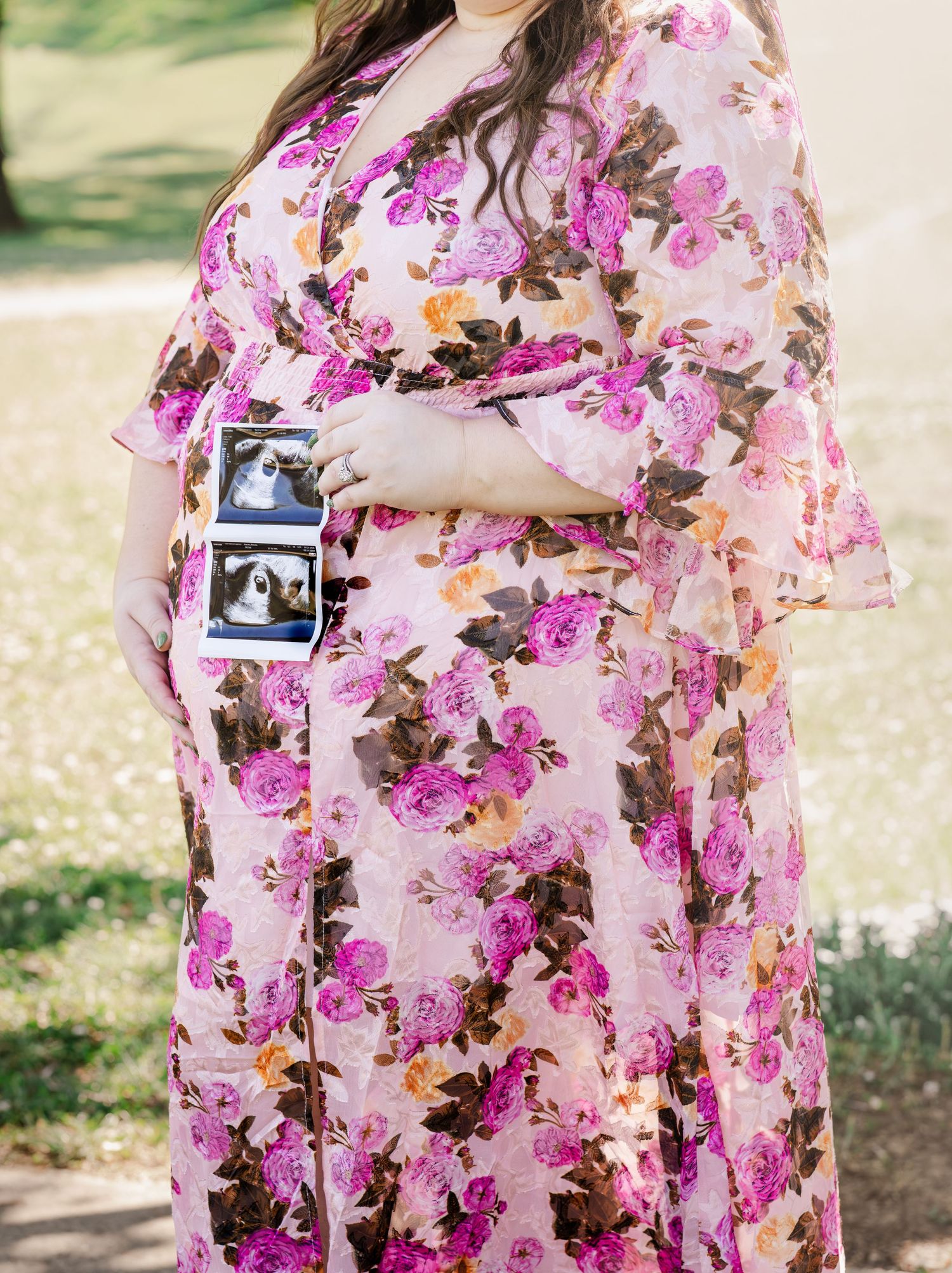 Pregnant woman in floral pink dress holding ultrasound photos outdoors during maternity photo session in Murfreesboro, Tennessee.