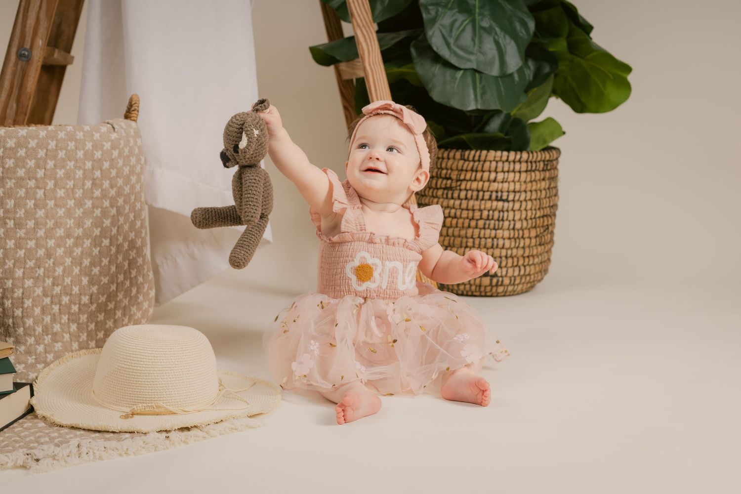 Baby girl in pink floral dress holds stuffed bunny toy, sitting near wicker basket and potted plant at a studio in Murfreesboro, Tennessee.