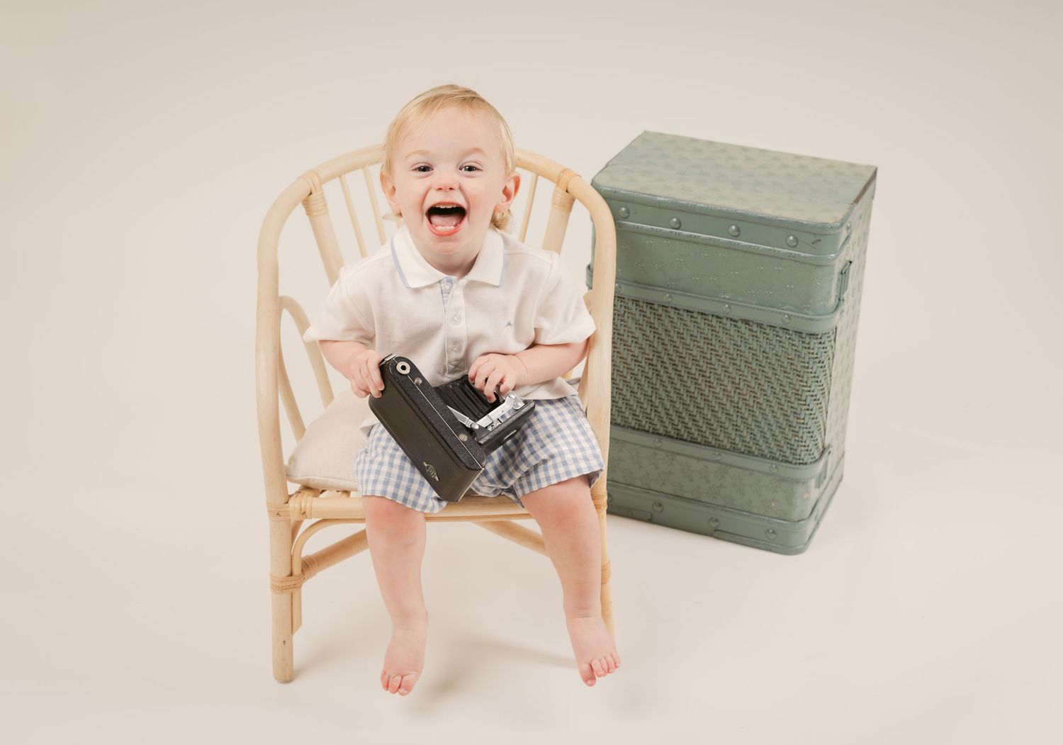 Happy toddler sitting in a wooden chair holding a vintage camera, next to green stacked boxes on a neutral background in Murfreesboro.