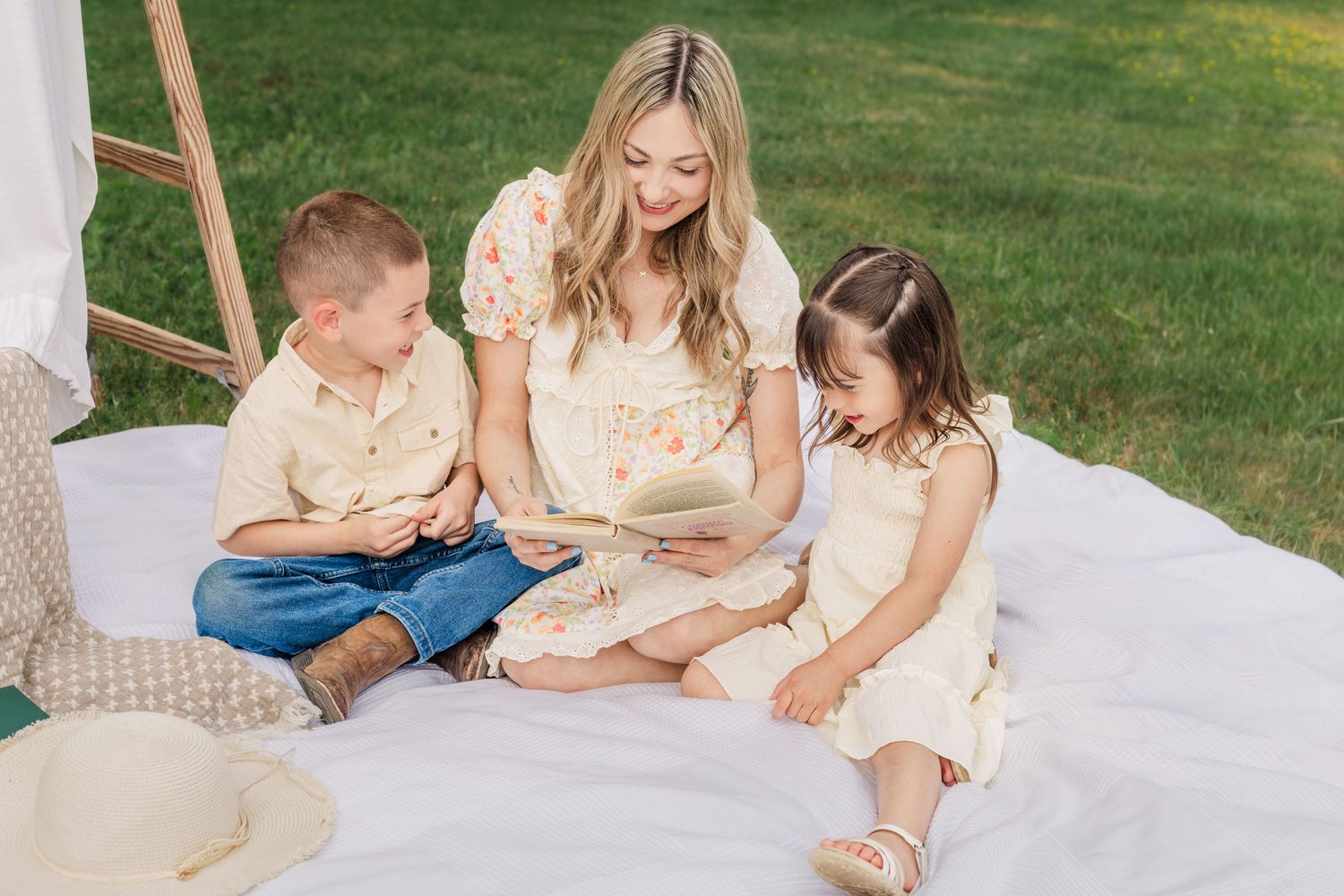 Woman reading a book with two young children on a white blanket outdoors in a lush green garden in Rutherford County.