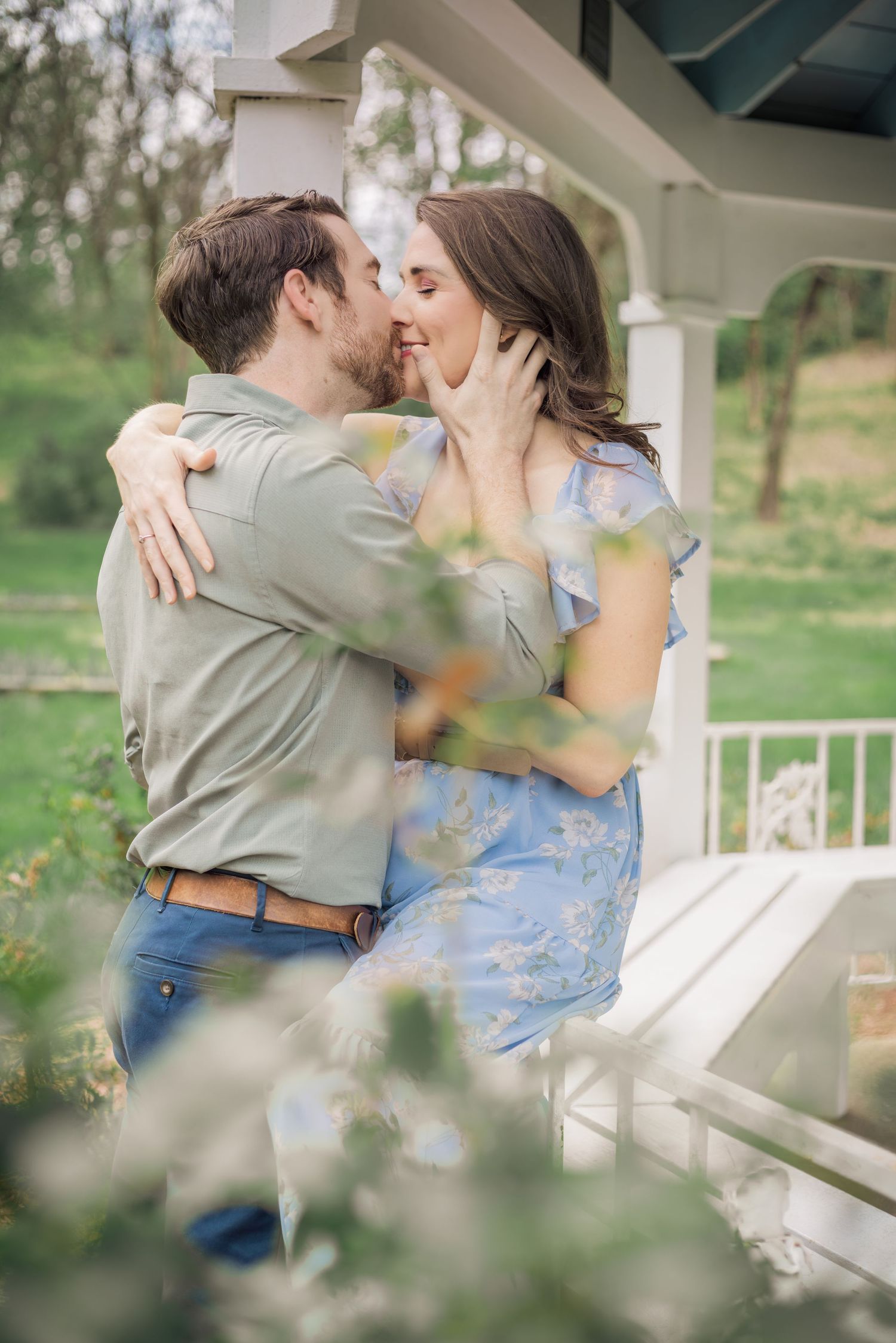 A couple shares a romantic kiss on a porch surrounded by greenery, woman in blue floral dress embracing man in sage shirt in Murfreesboro