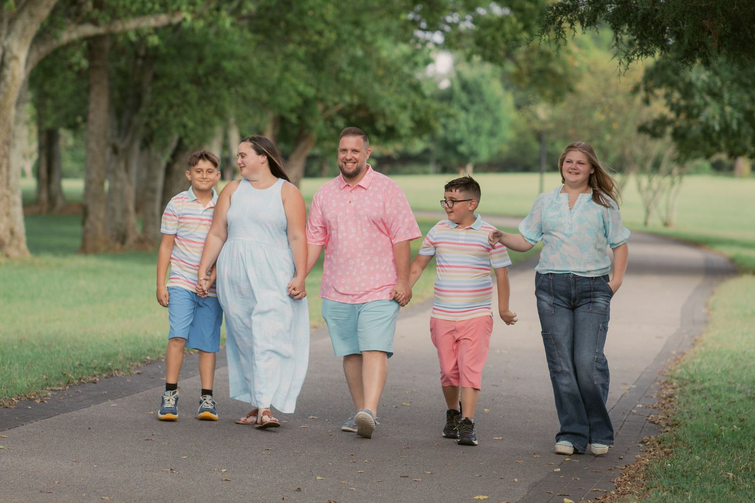 Family of five walking hand-in-hand on a park path surrounded by lush green trees on a sunny day in Murfreesboro, Tennessee.