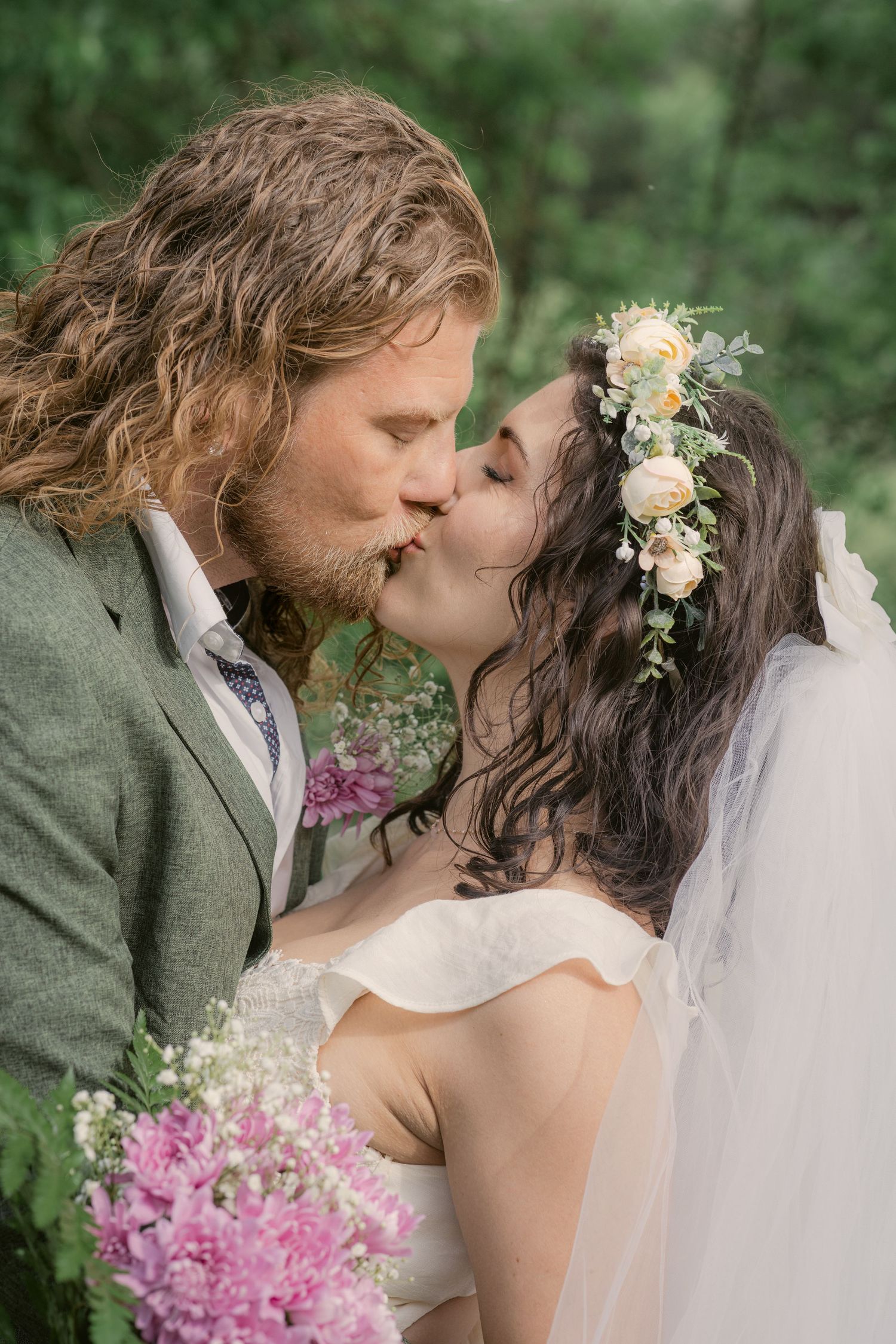 Bride and groom sharing a romantic kiss outdoors, bride wearing floral crown and veil, groom in gray suit holding pink flowers.
