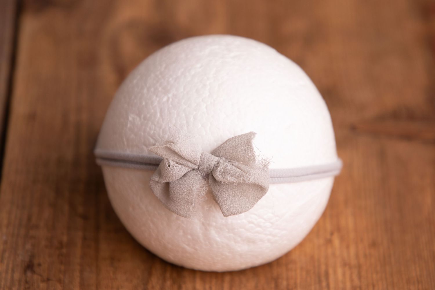 White fluffy newborn photography prop ball with a small gray bow headband on wood background.