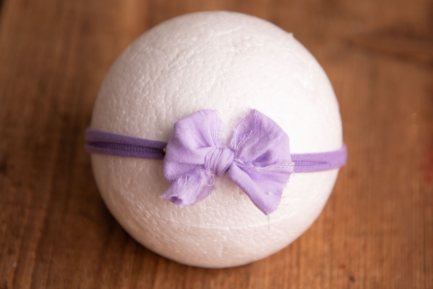 White knit ball with a purple fabric knotted bow headband on a wooden surface.