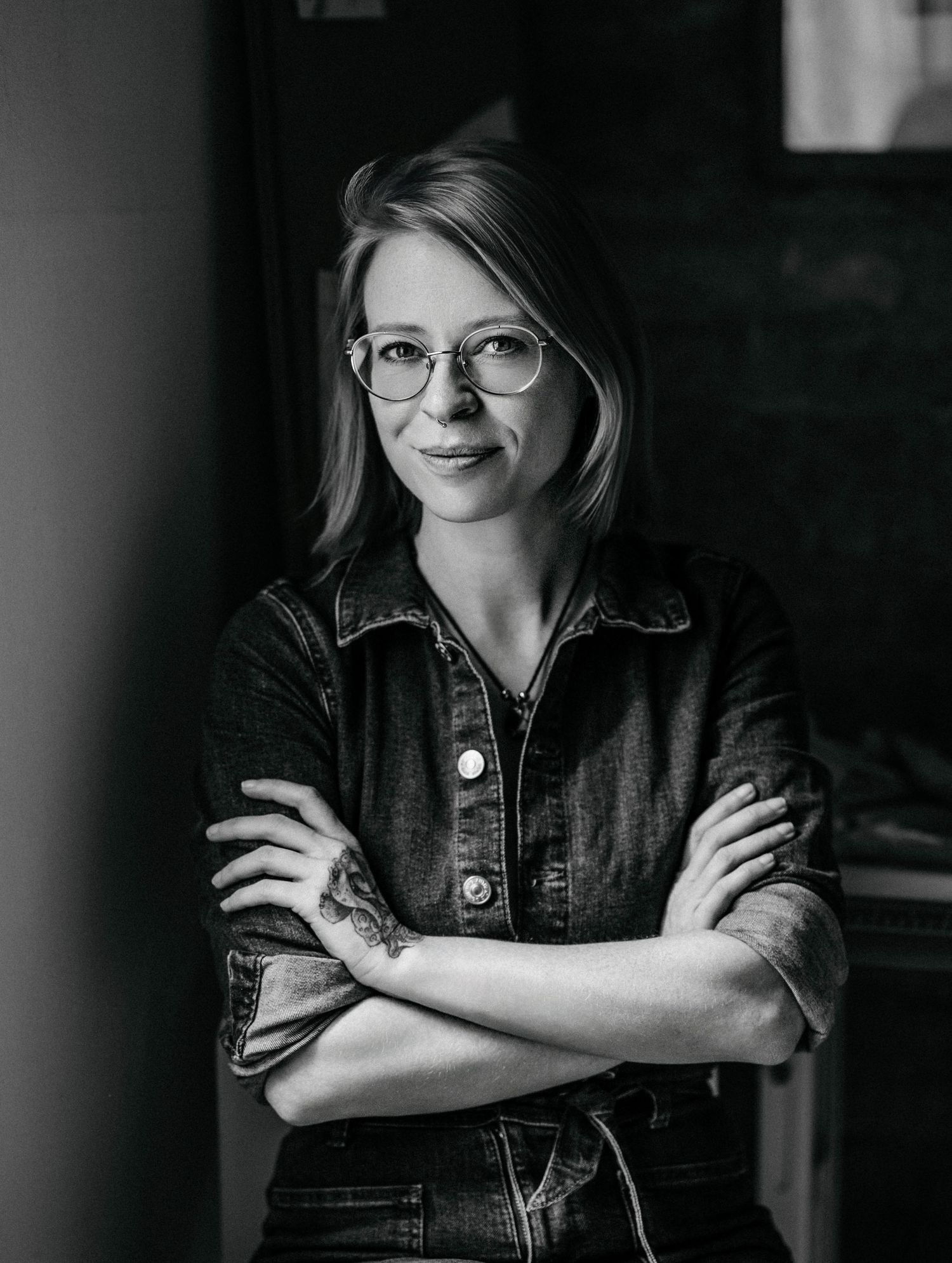 Black and white portrait of a young woman with glasses and crossed arms wearing a denim jacket with a visible tattoo.