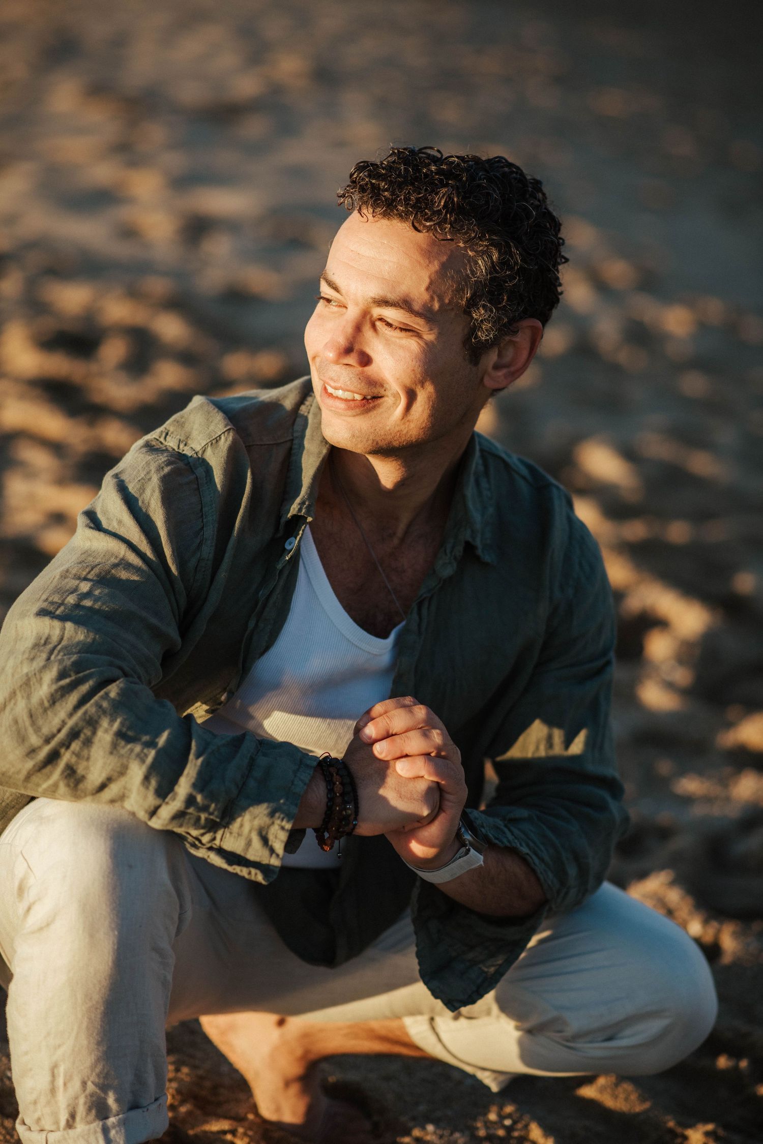 A person in a denim jacket sits cross-legged on the beach during golden hour, smiling warmly in the sunlight.