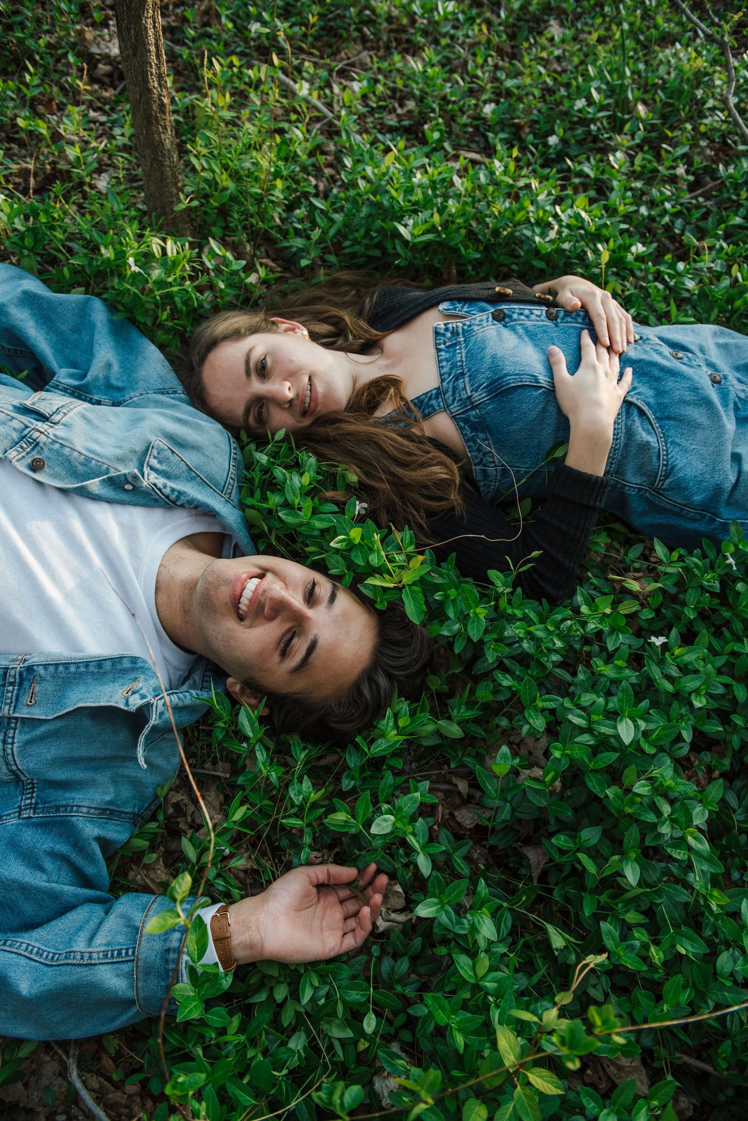Two people lying in green foliage wearing denim clothing while relaxing outdoors.