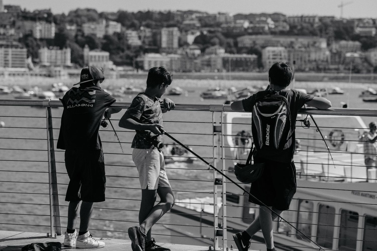 Three people stand along a waterfront railing overlooking a coastal cityscape in black and white photography.