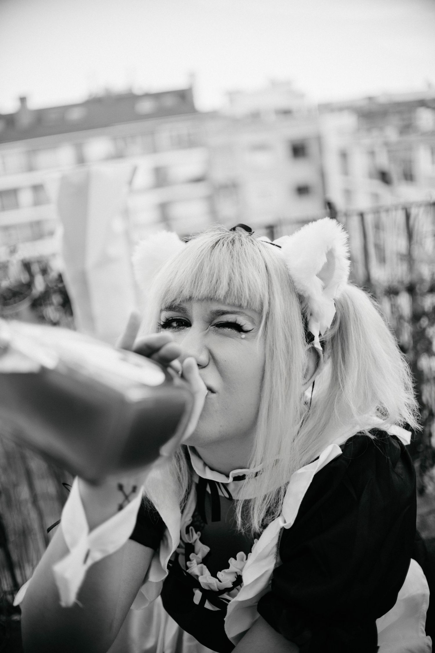 Black and white photo of a woman in anime maid cosplay with cat ears and blonde twintails drinking from a bottle outdoors.