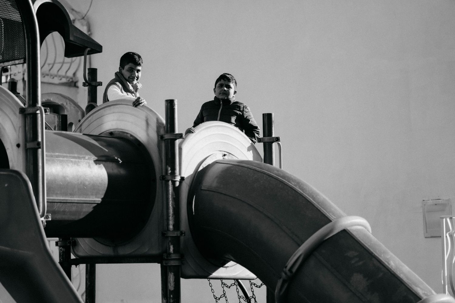 Black and white photograph of people playing on curved metal playground slides in a vintage setting.