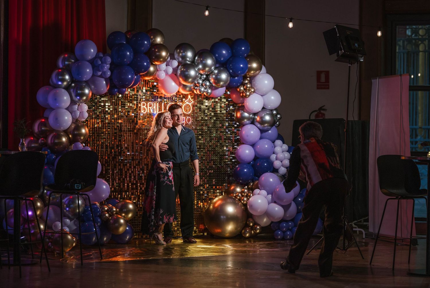 Couple posing at a festive balloon arch backdrop with purple and silver balloons at an indoor celebration event.