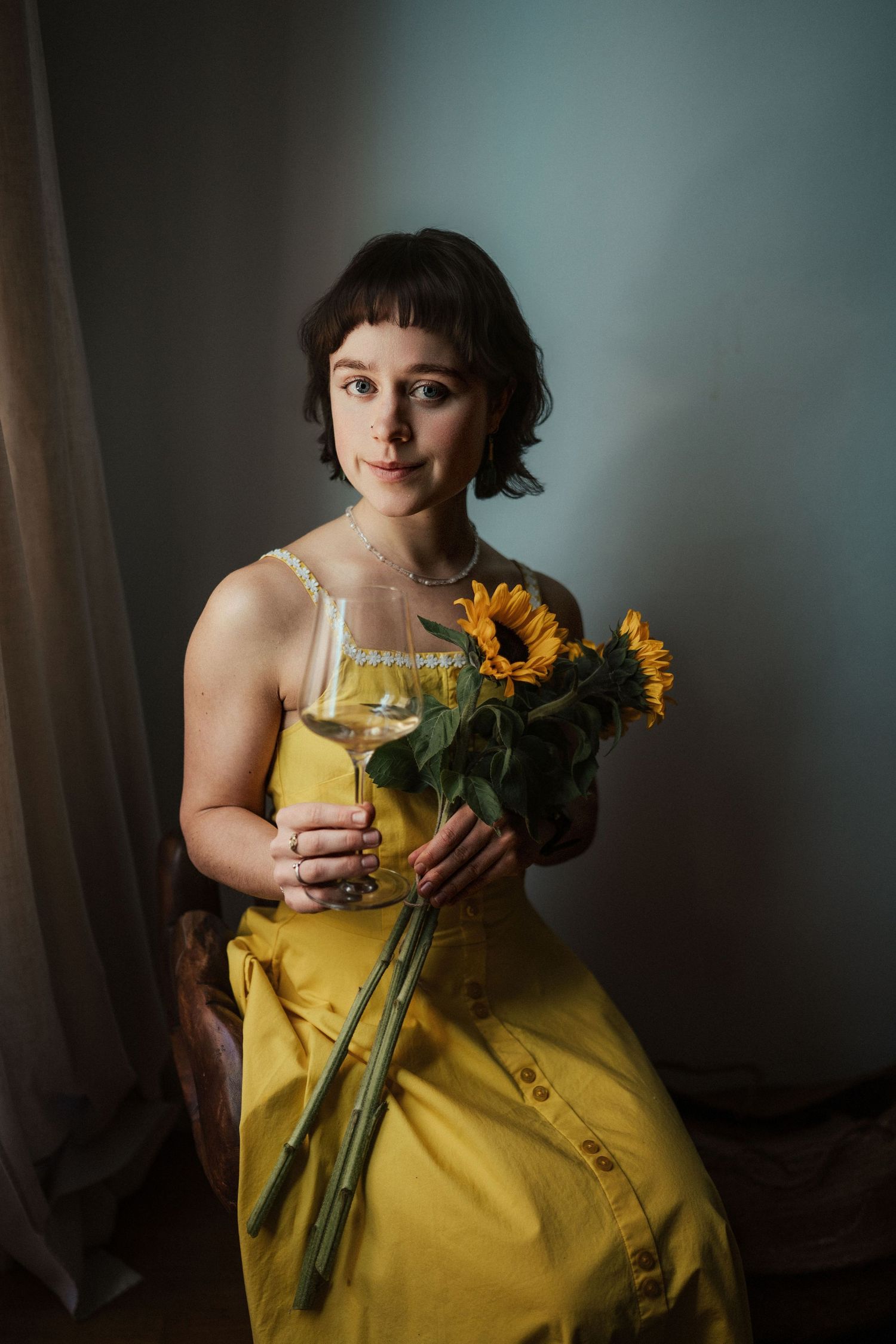 Young woman in yellow dress holding sunflowers and white wine glass, posed against gray wall.