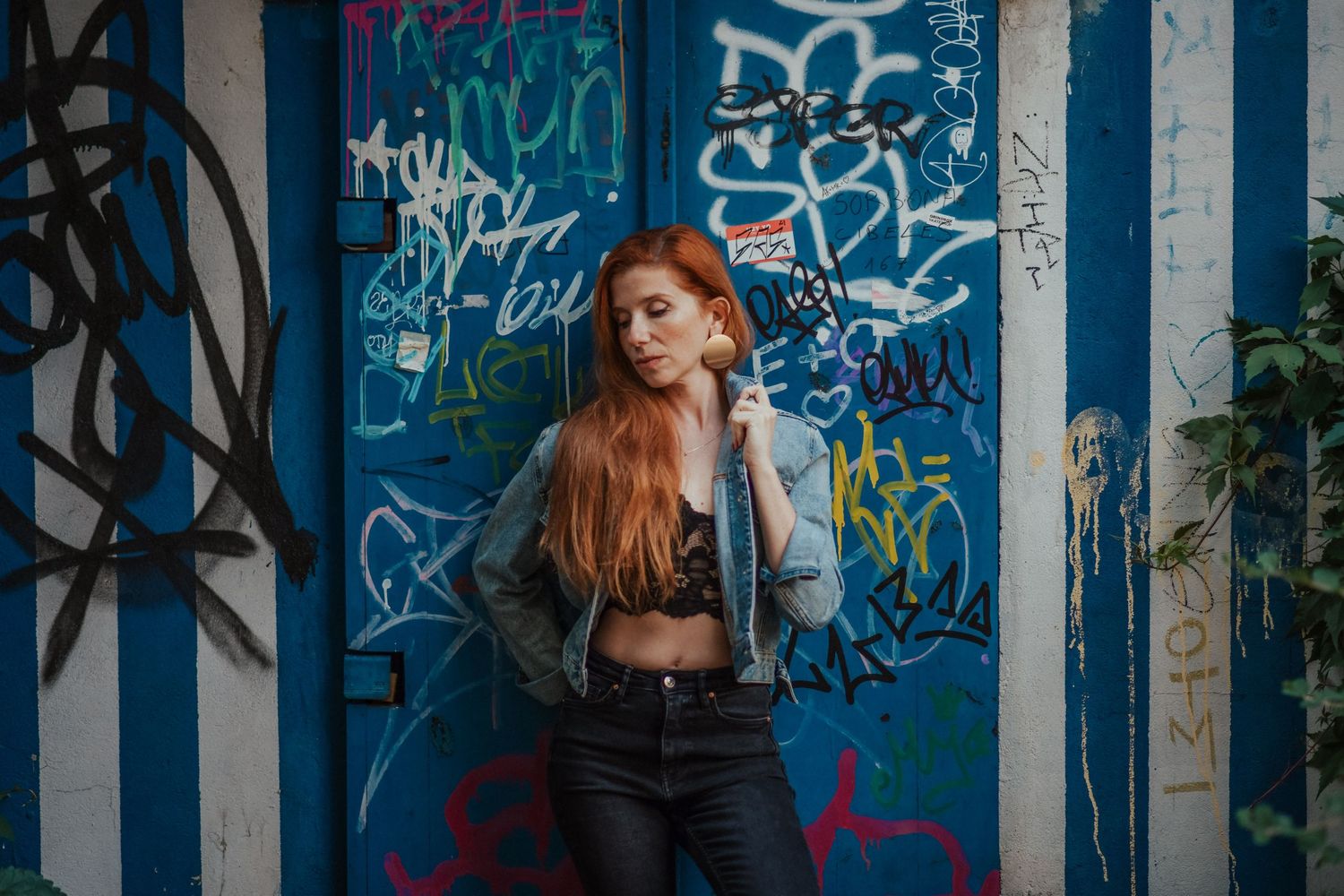 A person with red hair poses against a graffiti-covered wall with blue stripes in an urban setting.