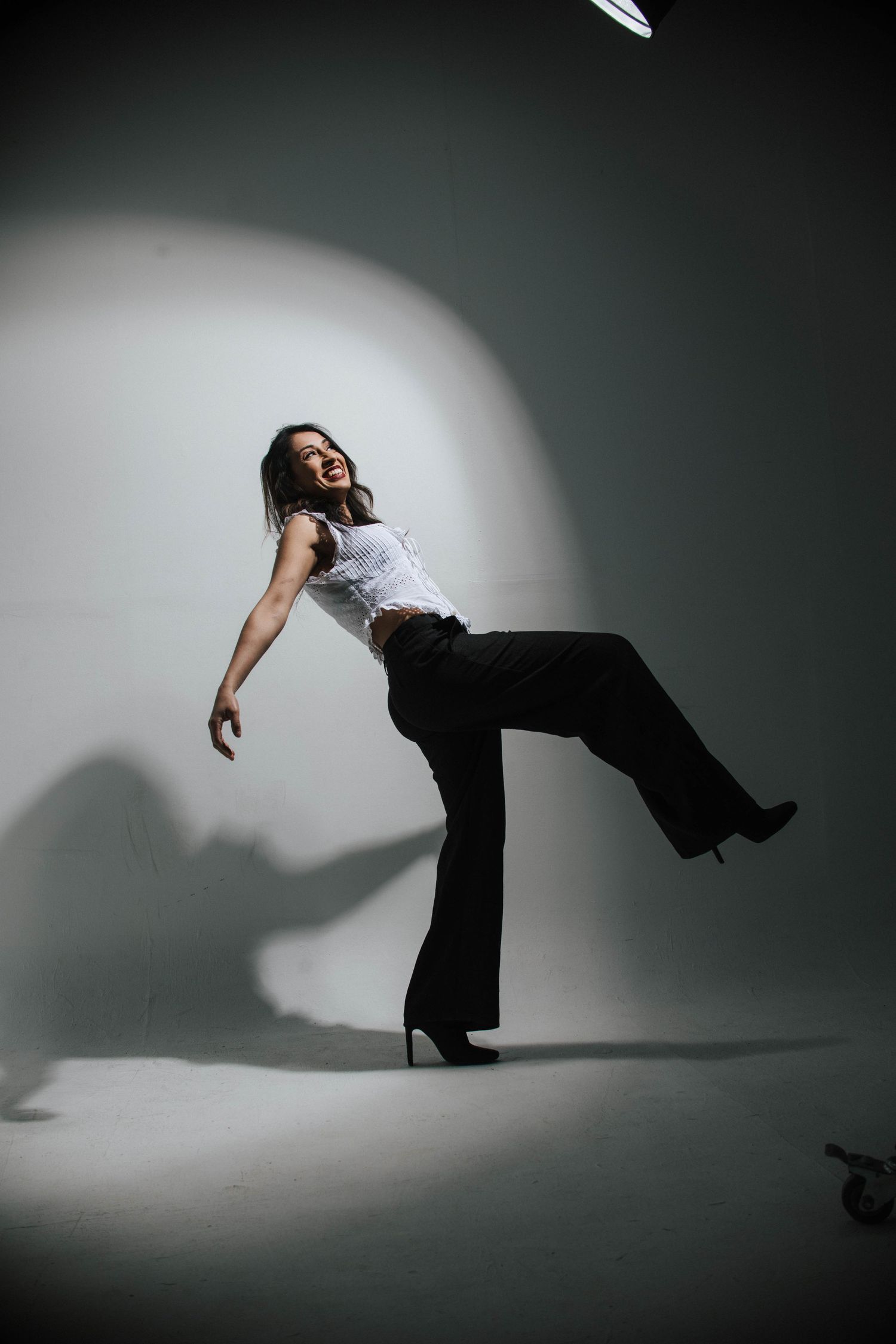Woman in white top and black flared pants poses dynamically in heels under studio spotlight.