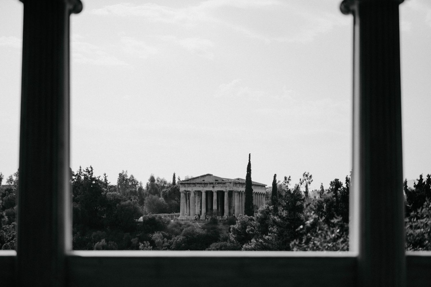 Black and white architectural photograph showing pillars and columns against a cloudy sky with trees in the background.