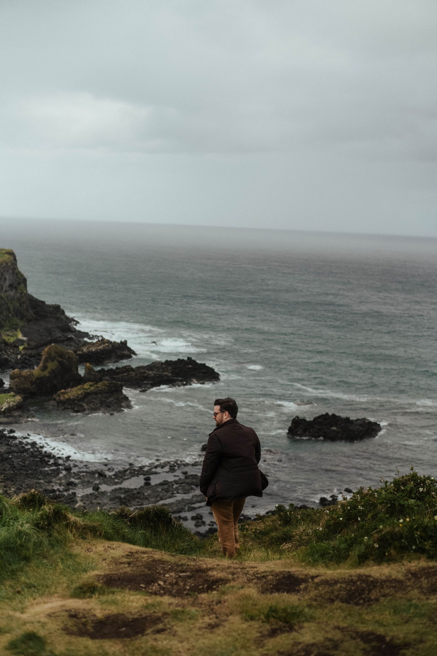 A moody image shows a man sitting on coastal cliffs overlooking a dramatic rocky shoreline and gray ocean waves.