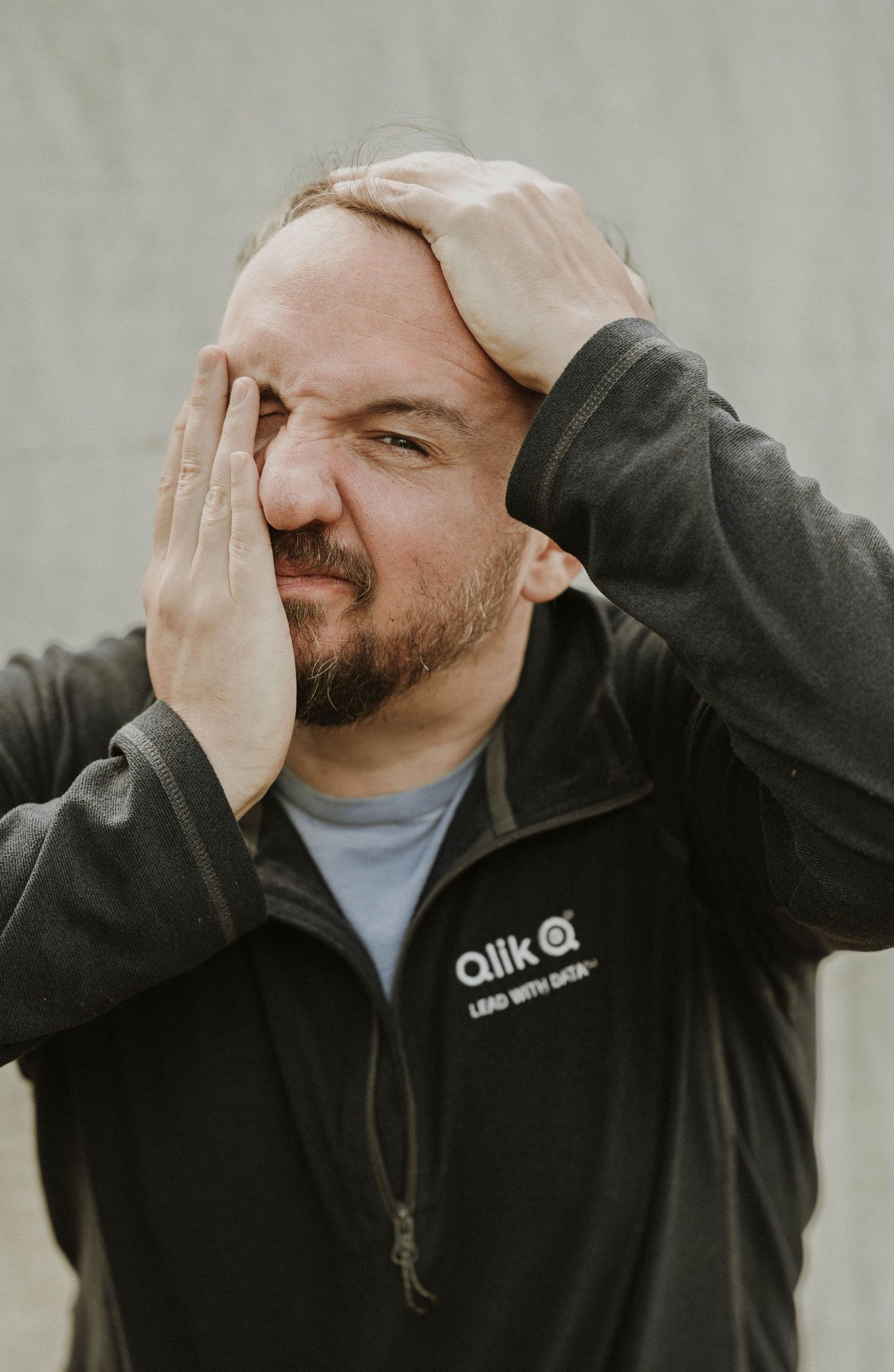 Stressed man in Qlik jacket holds head in hands with eyes closed, expressing frustration or overwhelm.