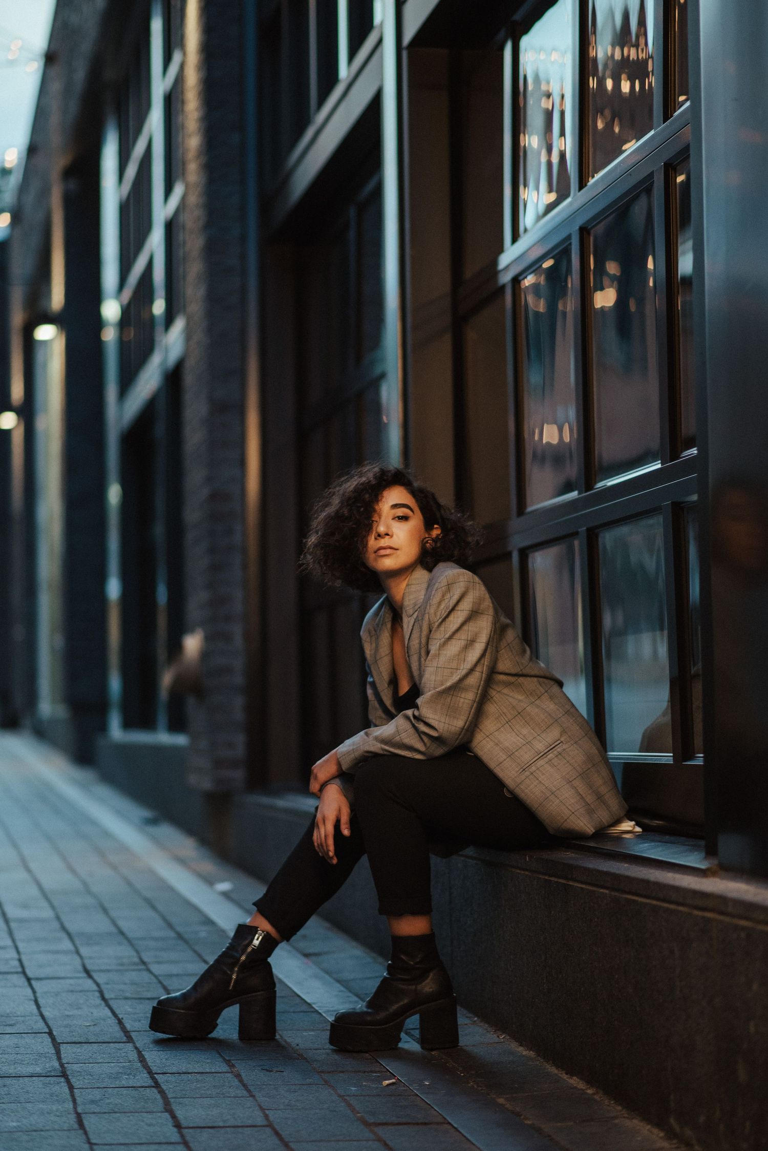 A person in a beige coat poses dramatically against a dark storefront window at night.
