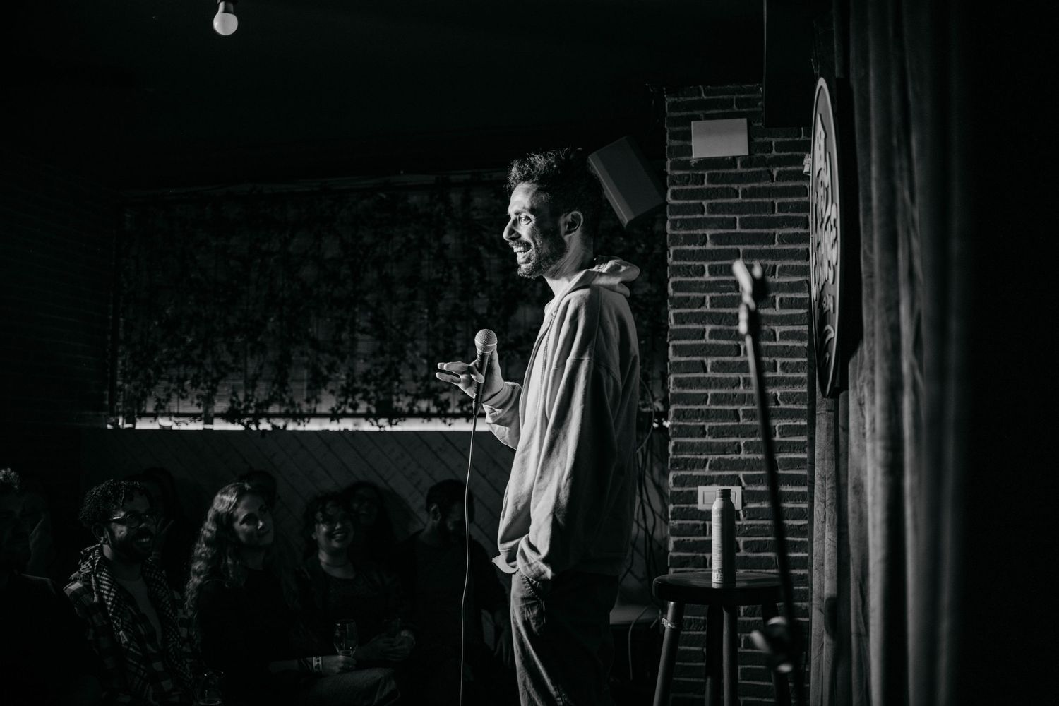 A performer stands on stage in dramatic black and white lighting at a comedy club.