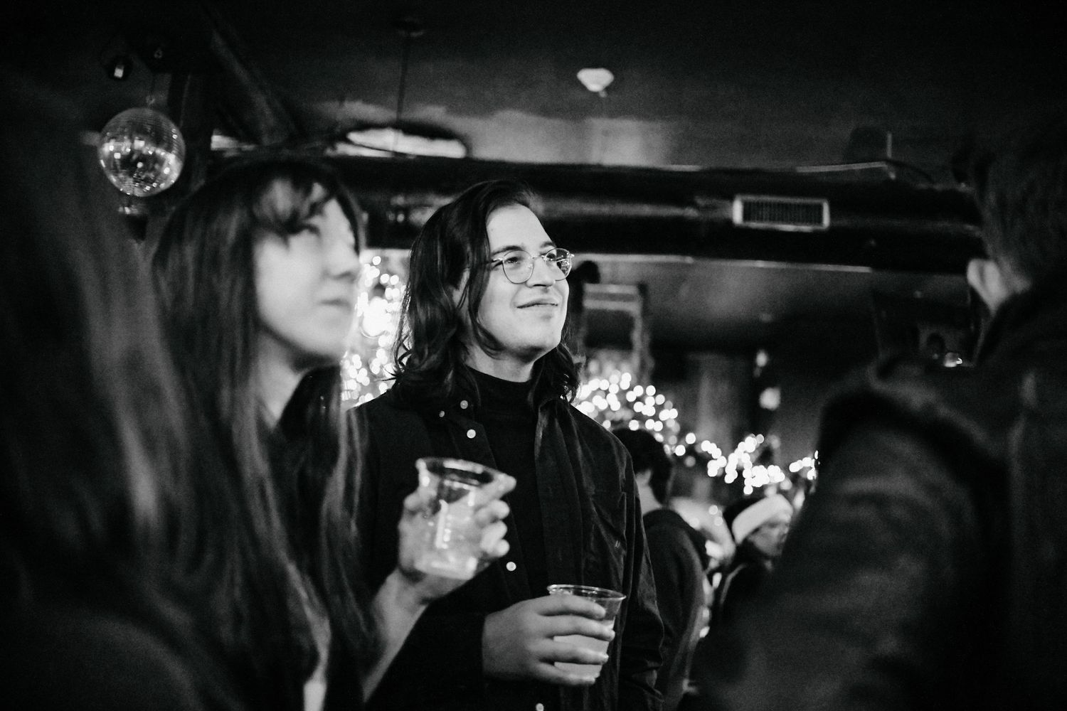 A black and white photo of people enjoying drinks and socializing at a dimly lit bar with festive lights in the background.