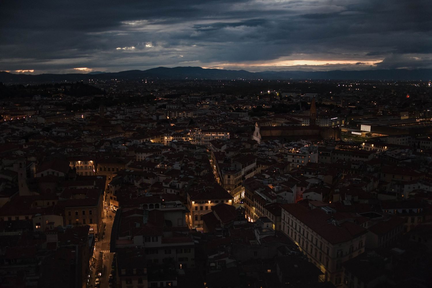 Panoramic aerial view of a city skyline at dusk with illuminated buildings and dark cloudy skies.