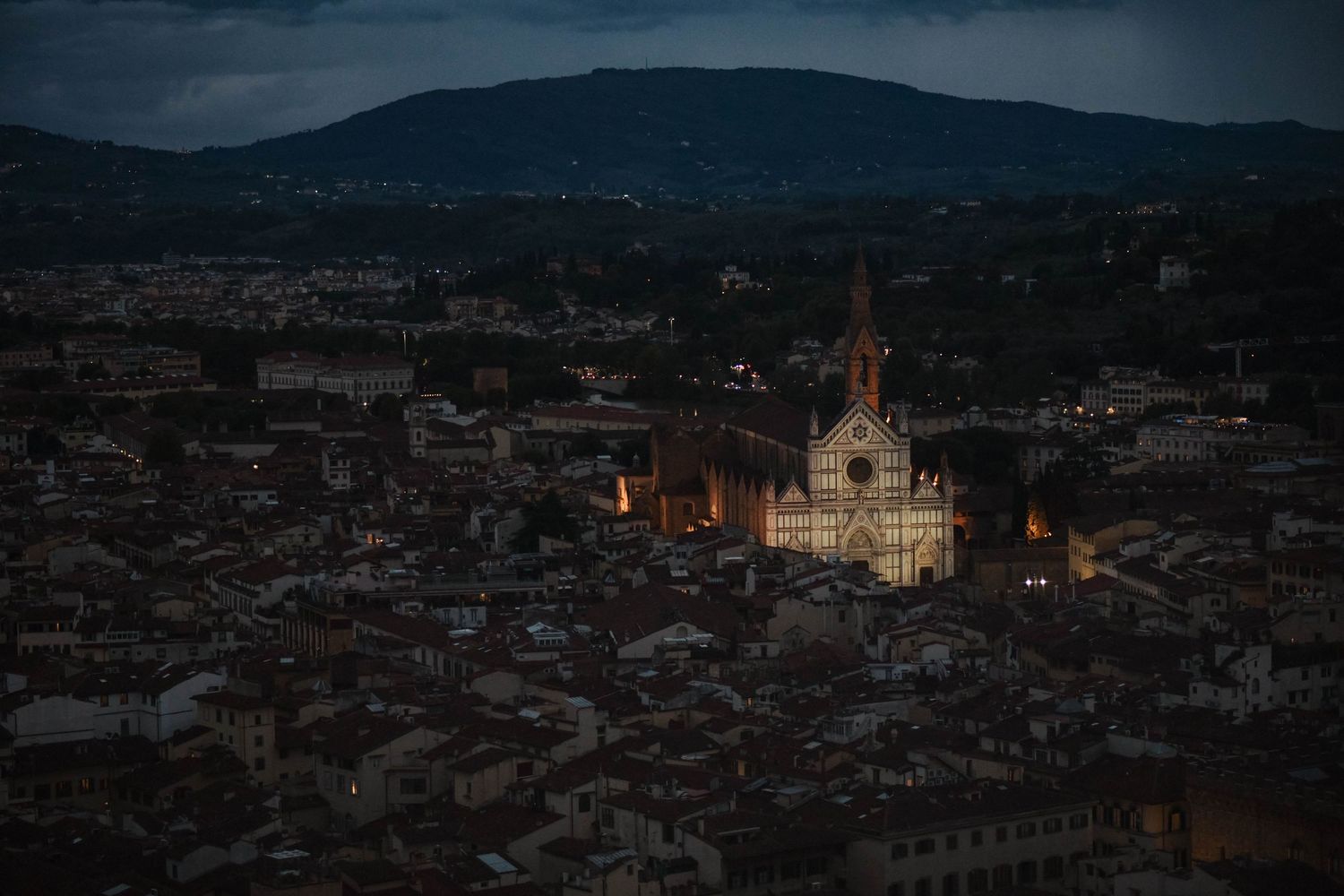 Panoramic aerial view of a historic European city at dusk with illuminated buildings and church spires dotting the skyline.