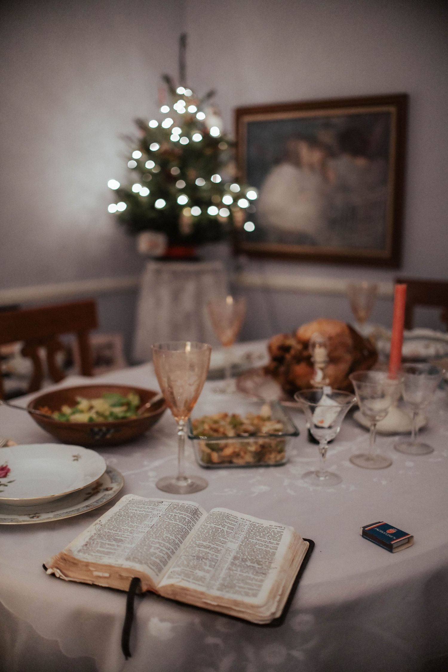 A festive Christmas dinner table setting with an open Bible, wine glasses, and a decorated tree in the background.