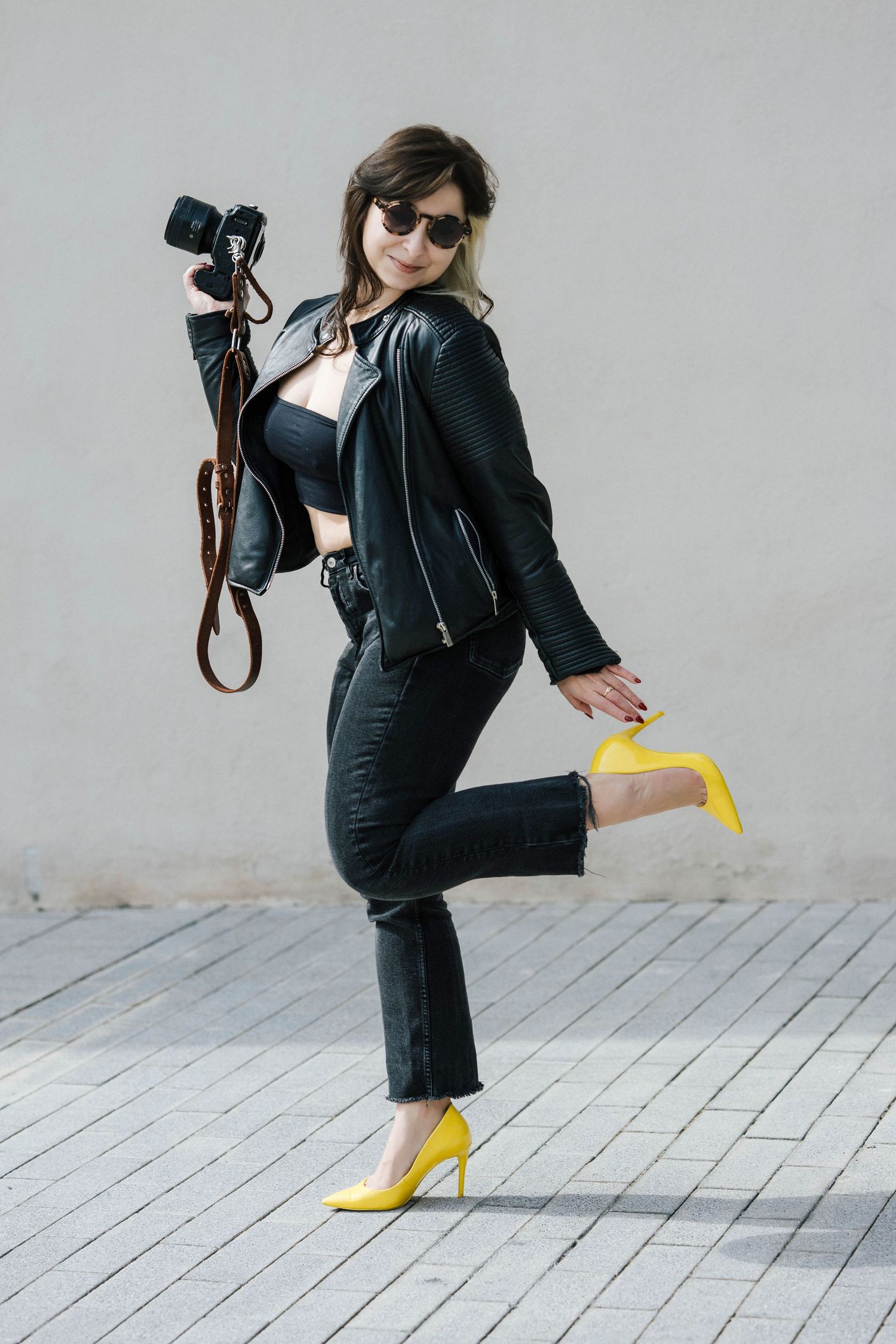 Photographer in black leather jacket and jeans strikes playful pose wearing bright yellow heels while holding camera.